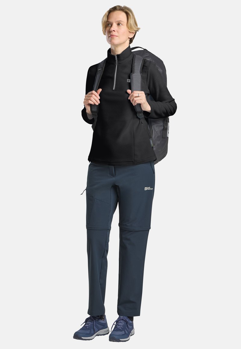 Pullover in pile nero con mezza zip, abbinato a pantaloni softshell blu. L'outfit è completato da uno zaino grigio e scarpe da trekking blu.