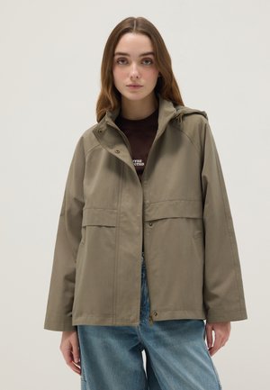 Jonge vrouw die een kaki capuchonjas draagt over een donkerbruin shirt en losse blauwe spijkerbroek, staand tegen een effen lichte achtergrond.