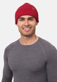 Red knitted beanie with a ribbed pattern and a small brown logo patch, worn by a male model in a grey long-sleeve shirt.