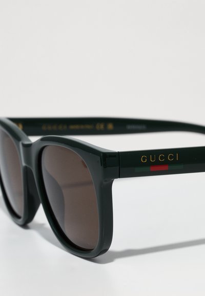 Groene plastic zonnebril met bruine lenzen. Gouden letters "GUCCI" op de zijkanten, verfraaid met rode en groene horizontale strepen. Rechthoekige vorm.