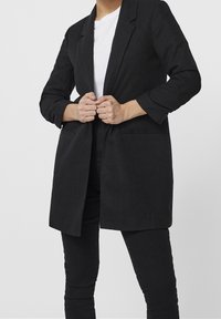 Blazer noir texturé à manches longues, col cranté et devant ouvert. Dispose de deux poches et est porté sur une chemise blanche et un pantalon noir.