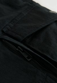 Gros plan sur une fermeture éclair noire partiellement ouverte sur un tissu noir texturé avec des coutures visibles et un bouton à proximité.