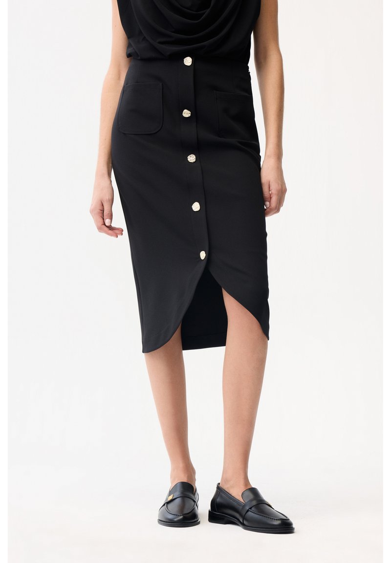 Frau trägt einen schwarzen, geknöpften Midirock mit Fronttaschen und schwarze Slip-On-Loafer, steht vor einem weißen Hintergrund.