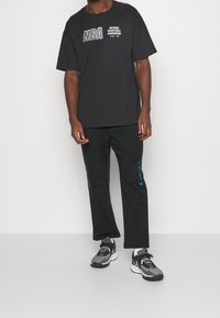 Schwarzes NBA-Graphic-T-Shirt mit weißer Schrift, kombiniert mit schwarzen Jogginghosen mit türkisfarbenen Akzenten. Trägt dazu schwarze und graue Sneakers.