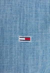 Jasnoniebieska tkanina o fakturowanym splocie z prostokątnym haftowanym logo: czerwone, białe i czarne paski na białym tle.