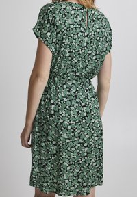 Vihreä kukkakuosinen mekko tummalla taustalla, lyhyet hihat, vyötärö nidottu ja pieni halkio takakauluksessa. Kevyt kangas, polvipituinen.