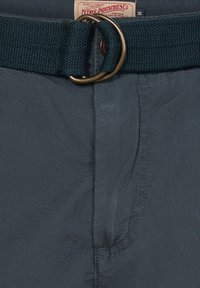 Elastiske shorts i mørkeblå stof. Har et struktureret bælte med en bronze metalring og et mærke på taljen.