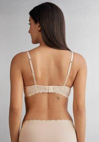 Beige bh med spetsbak och justerbara band, med en slät rem och scalloped spetsbård. Inkluderar en mittknutdetalj.