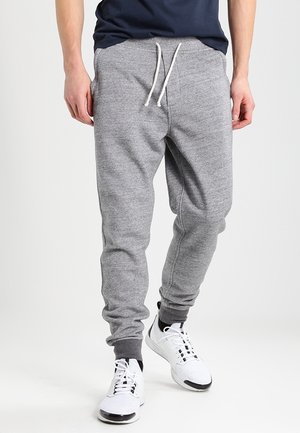 Homme portant un pantalon de survêtement gris avec cordon de serrage et des baskets blanches, debout devant un fond uni.