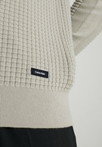 Hellgrau strukturiertes Strickpullover mit geripptem Saum, ausgestattet mit einem schwarzen Calvin Klein Logo-Patch an der Seite.
