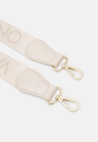 Béžový látkový popruh tašky s texturovanými koženými konci a zlatými otočnými sponami na bílém pozadí.