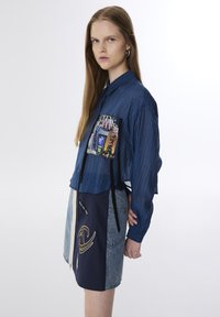 Denim overhemd met knopen in blauw, voorzien van een grafisch zakdetail; gecombineerd met een twee-kleurige rok met een rits en geborduurde accenten.