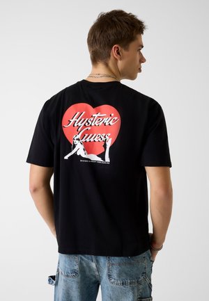 Junger Mann trägt ein schwarzes T-Shirt mit rotem Herz und weißem "Hysteric Guess"-Schriftzug sowie einer Grafik einer Frau auf dem Rücken, kombiniert mit blauen Jeans.