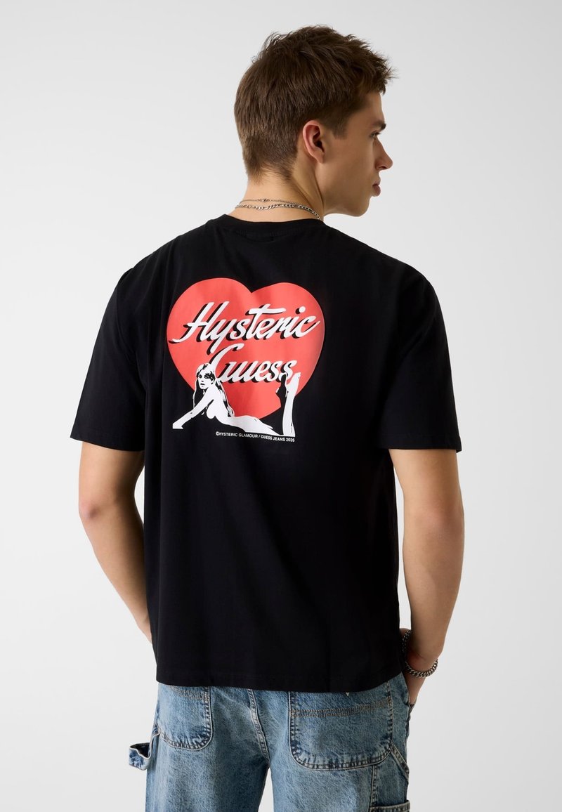 Νέος άνδρας που φοράει μαύρο T-shirt με κόκκινη καρδιά και λευκό κείμενο "Hysteric Guess" και γραφικό κάποιας γυναίκας στην πλάτη, συνδυασμένο με μπλε τζιν.