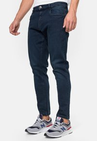 Donkerblauwe denim jeans met een slim fit, met een klassieke vijfzakkenontwerp en een kleine metalen knoopsluiting. Draag ze met grijze en marineblauwe sneakers.