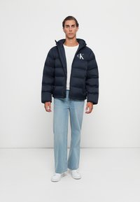 Man in een marineblauwe pufferjack met capuchon, een wit T-shirt, lichte blauwe jeans en witte sneakers, staand tegen een witte achtergrond.
