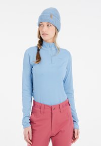 Maglietta azzurra chiara a maniche lunghe con colletto a mezza zip, abbinata a pantaloni da neve rosa con dettagli laterali e un berretto lavorato blu texturizzato.