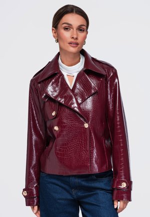 Donna che indossa una giacca di pelle lucida rosso scuro con texture coccodrillo sopra una maglia bianca e jeans blu, rivolta in avanti con espressione neutra.