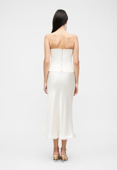 Γυναίκα με μακριά σκουρόχρωμα μαλλιά, φορώντας λευκό φόρεμα χωρίς τιράντες με φερμουάρ στην πλάτη και χρυσά ψηλοτάκουνα σανδάλια, στέκεται μπροστά σε απλό φόντο.