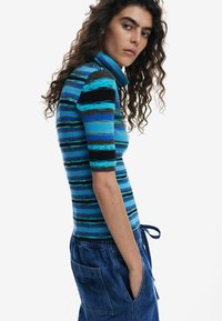 Fitted pullover with a high collar, featuring blue and black horizontal stripes. Made of textured knit material, paired with loose denim shorts.