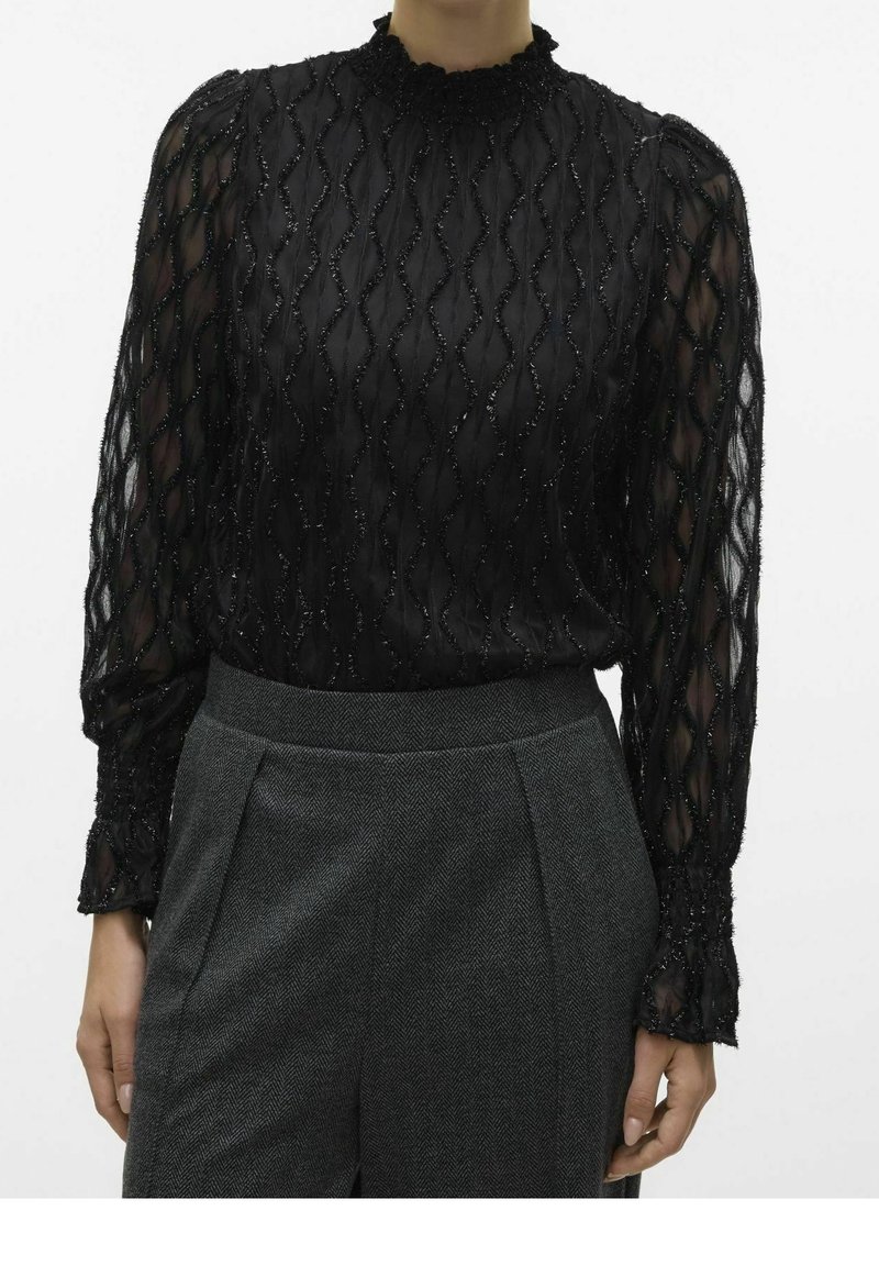 Blouse noire transparente avec motif ondulé texturé et manches longues, associée à un pantalon gris à chevrons taille haute.