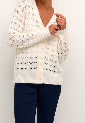 Personne portant un cardigan blanc en tricot boutonné avec un motif festonné et un pantalon bleu foncé, ajustant la manche avec la main.