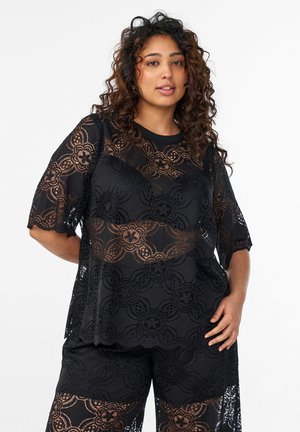 Kobieta o kręconych włosach, ubrana w czarny koronkowy top w kwiaty i pasujące koronkowe szorty, stoi z jedną ręką na biodrze na białym tle.