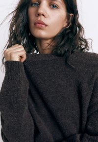 Giovane donna con capelli castano ondulati che indossa un maglione marrone scuro lavorato a maglia, tenendo il colletto vicino al volto su uno sfondo semplice.