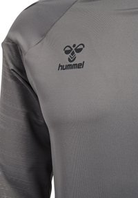 Nahaufnahme eines grauen Sportshirts mit schwarzem hummel-Logo auf der Brust und strukturierten, dunkler grauen Ärmeln.