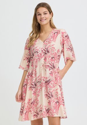 Femme souriante portant une robe crème longueur genou avec des motifs floraux cachemire rouges et noirs et des manches mi-longues, une main dans la poche.