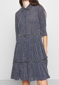 Mörkblå klänning i genomskinligt tyg, med ett prickigt mönster, knappstängning framtill, krage och ett flerlagers volangslut med trekvartslånga ärmar.