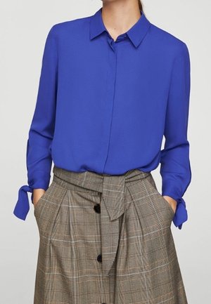 Kobieta w kobaltowo niebieskiej bluzce z długim rękawem z wiązanymi mankietami oraz brązowej, wysokojakościowej spódnicy w kratę z paskiem wiązanym z przodu i czarnymi guzikami.