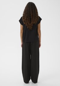 Donna con lunghi capelli ricci che sta in piedi di spalle, indossando una maglietta nera a maniche corte e pantaloni neri a gamba larga su uno sfondo uniforme.