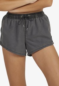 Naine kannab tumehallide sportlike lühikesi pükse, millel on elastne vöökoht ja must nöör, seisab käed üle palja kere risti.