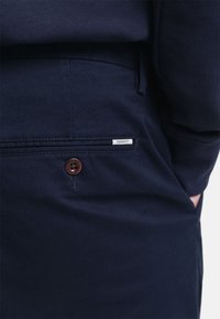 Navyblaue Hose aus strukturiertem Stoff mit einer Seitentasche, ausgestattet mit einem braunen Knopf und einem kleinen weißen Etikett mit dem "GANT"-Logo.