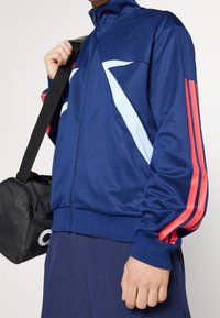 Person wearing blue track jacket with light blue and red stripes, holding black shoulder bag with white logo.
