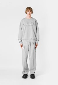 Sudadera gris con logo en relieve, combinada con pantalones deportivos grises que tienen franjas laterales y bolsillos. El modelo lleva zapatos negros tipo slip-on.