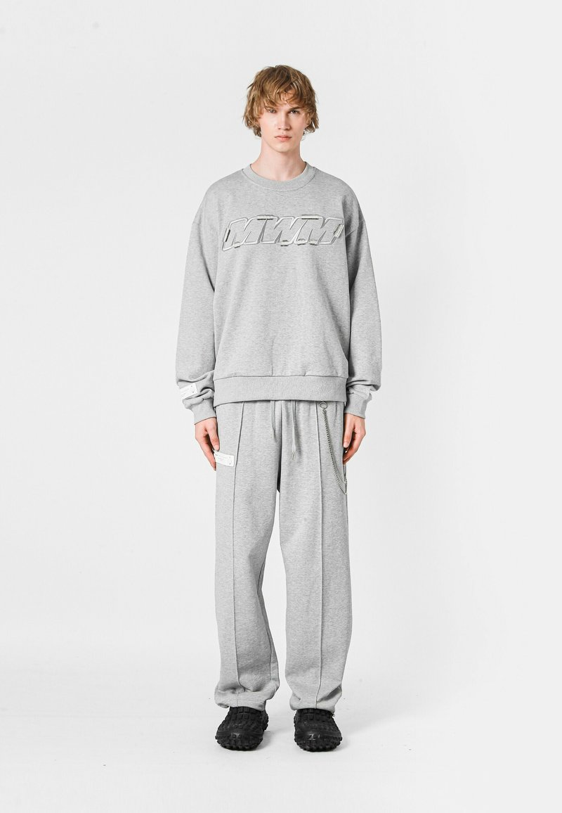 Sudadera gris con logo en relieve, combinada con pantalones deportivos grises que tienen franjas laterales y bolsillos. El modelo lleva zapatos negros tipo slip-on.