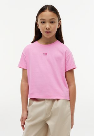 Νεαρό κορίτσι με μακριά σκούρα μαλλιά που φοράει ένα ροζ κοντό μπλουζάκι και μπεζ παντελόνια, στέκεται μπροστά σε λευκό φόντο.