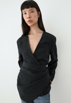 Femme aux longs cheveux foncés portant un chemisier noir style cache-cœur avec un décolleté en V profond et un jean bleu, debout devant un fond clair uni.