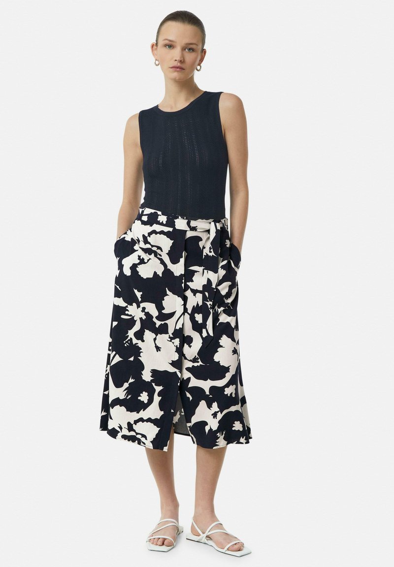 Marineblaues Stricktanktop, kombiniert mit einem midi Rock mit weiß-blauem Blumenmuster, der Taschen und einen Bindegürtel hat. Weiße, gepolsterte Sandalen mit Riemchen.