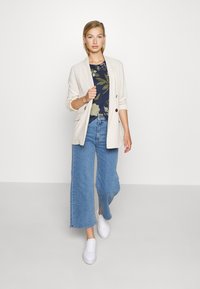 Blazer de linho bege claro sobre uma blusa floral escura, combinado com jeans de perna larga azuis e ténis brancos. Textura suave, ajuste descontraído.