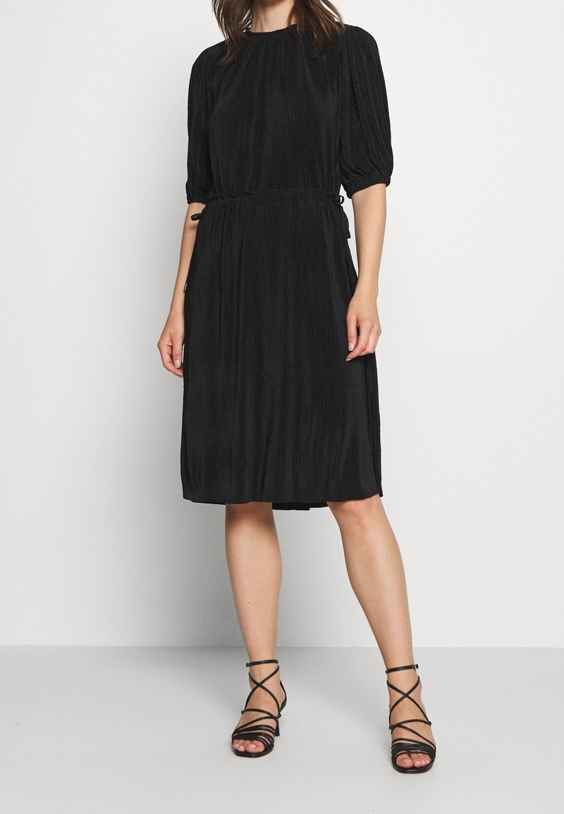 Femme portant une robe noire mi-longue à manches bouffantes et des sandales à talons hauts noires à brides, debout contre un mur blanc.
