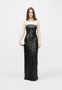 Juoda blizgučių suknelė be petnešėlių, aptemptos silueto, iki grindų ilgio ir švelnios tekstūros. Papildyta maža balta delninuke.