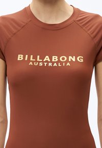 Közelkép egy személyről, aki rozsdaszínű Billabong Australia rövid ujjú inget visel, varrott vállvarrással.