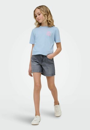 Meisje loopt en draagt een lichtblauw T-shirt met "HEY YOU!", grijze spijkershorts en witte sneakers tegen een effen lichte achtergrond.