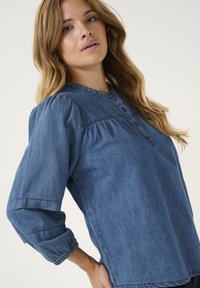 Denimová blůza ve středně modré barvě, se zaobleným límcem, knoflíkovou légou a nařasenými rukávy se pružnými manžetami. Měkká, strukturovaná tkanina.