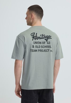 Homme de profil gauche montrant le dos d'un T-shirt gris avec le texte noir « Heritage Union of '63 & Old School Team Project Inc. »