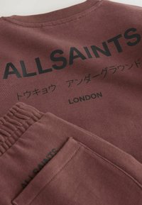 Nærbillede af et brunt tøjstykke med sort tekst "ALLSAINTS," japanske tegn og "LONDON," der viser ribbet krave og elastisk talje.