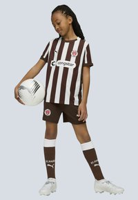 Brun och vit randig fotbollsjersey med logotyp, matchande shorts, vita strumpor med bruna accenter, håller i en grå och vit fotboll.